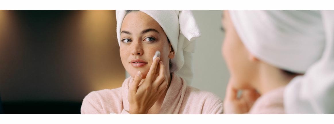 Woman with a towel on her head applies cream to her face with her hands
