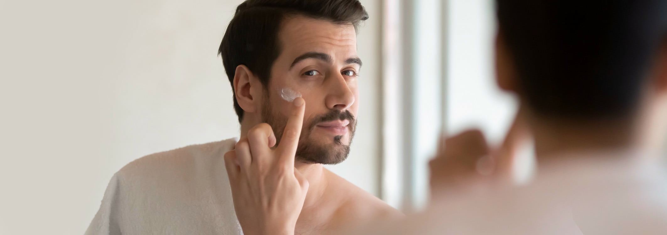 mann welcher in den spiegel schaut und sich mit dem zeigefinger creme auf die wangen schmiert