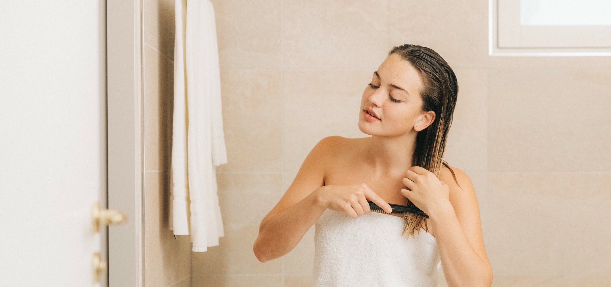 frau sitzt in badetuch auf der badewanne und kämmt sich die nassen haare
