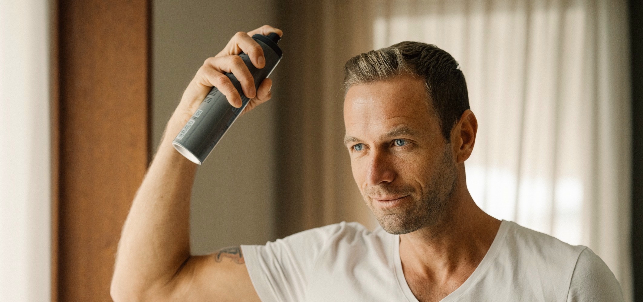mann sprayt sich mit grauer dose haarspray auf hell braunes kurzes haar