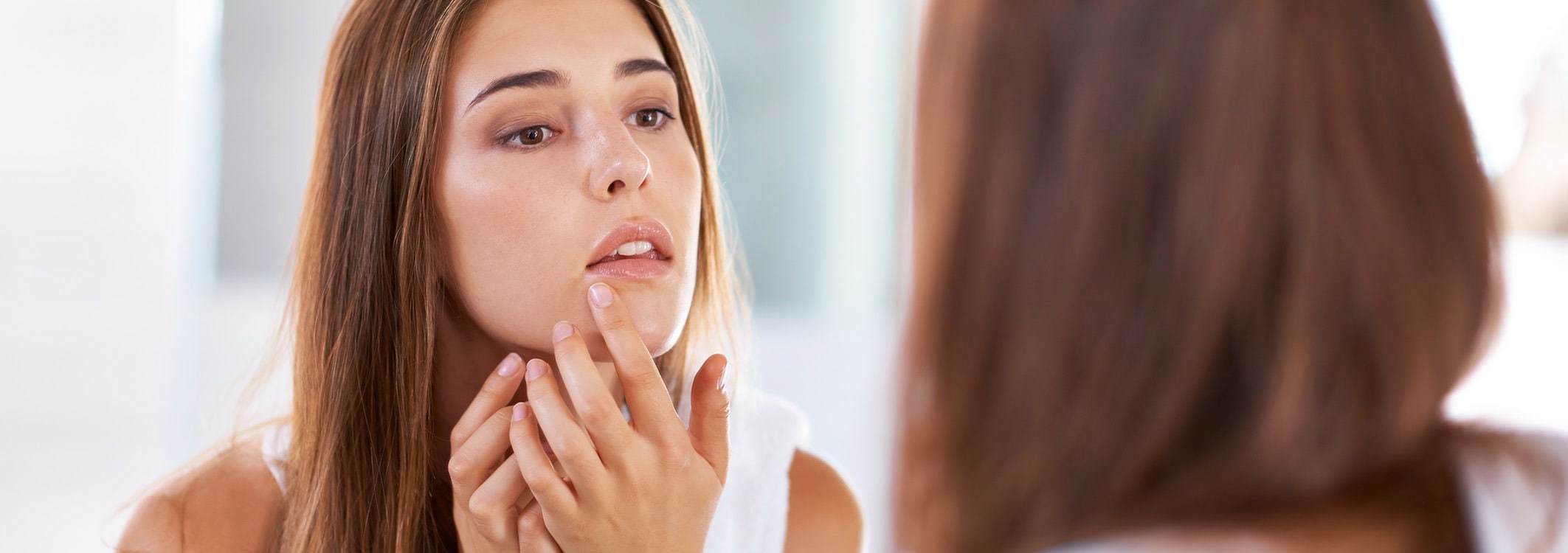 Brown-haired woman looking in the mirror and searching for spots on her chin with her fingers